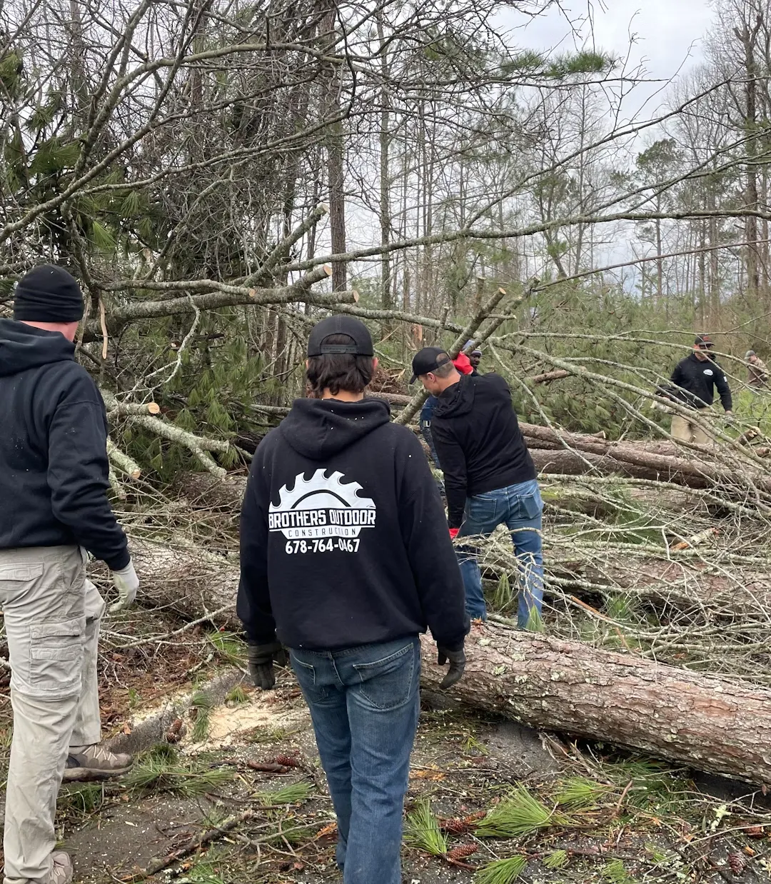 about-1 Four people in outdoor workwear clearing fallen trees and branches in a wooded area after a storm.