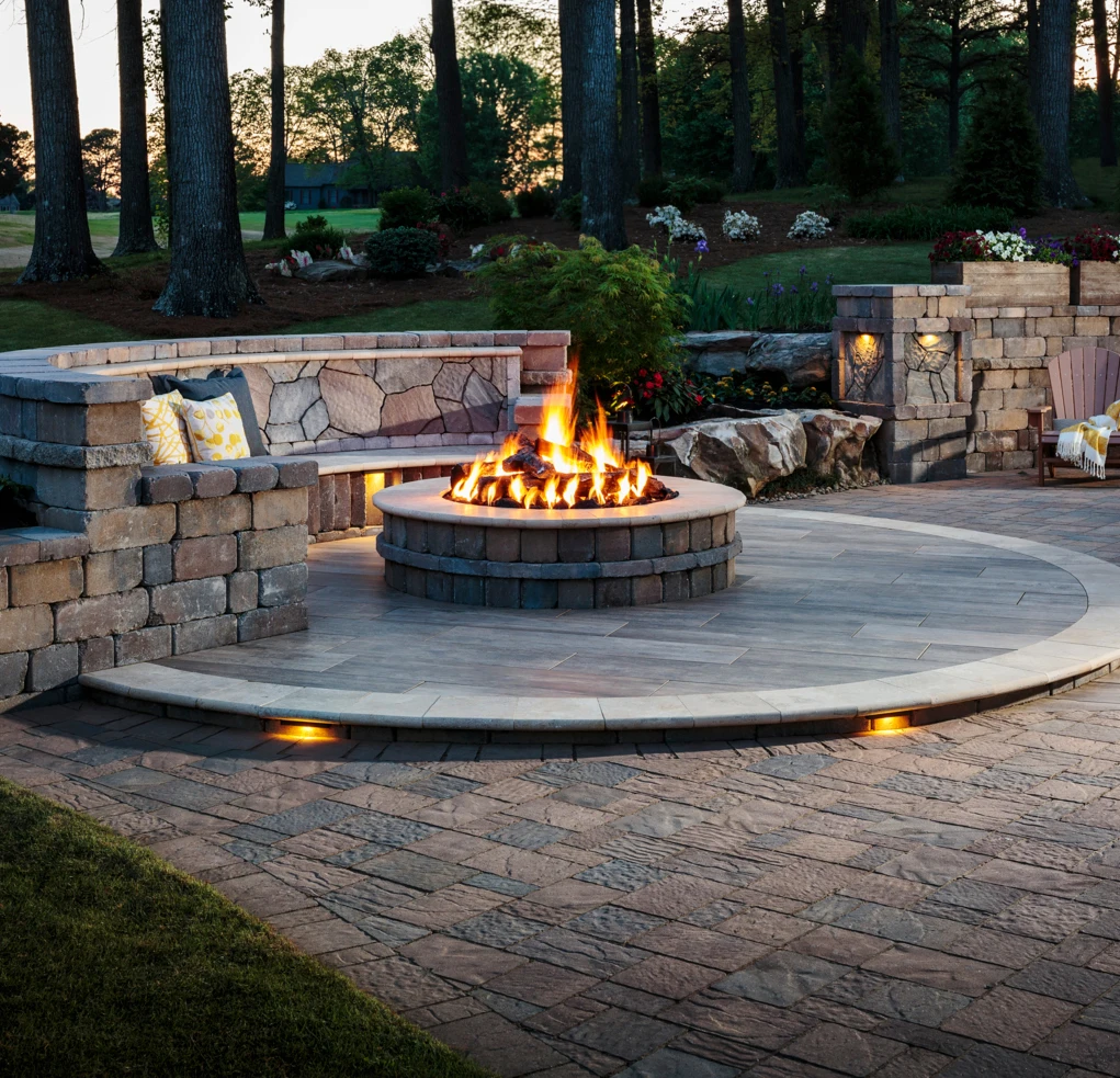 fire-pit-1 Stone patio with a round fire pit, built-in benches, glowing lights, and trees in the background at dusk.