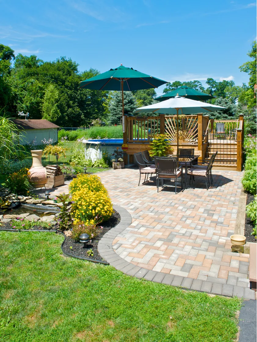 landscaping-2 Sunny patio with a table and chairs, large umbrellas, garden, and a small pond in a backyard setting.