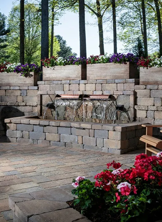 water-3 Stone patio with a tiered brick water fountain, colorful flowers, and tall trees in the background.