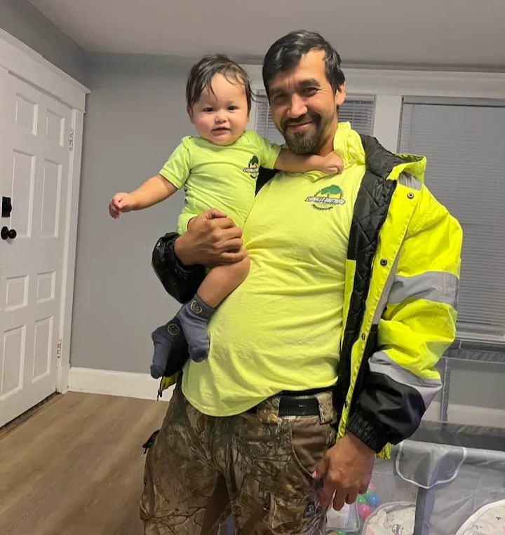 A smiling man holds a toddler; both wear matching green shirts and stand in a living room.